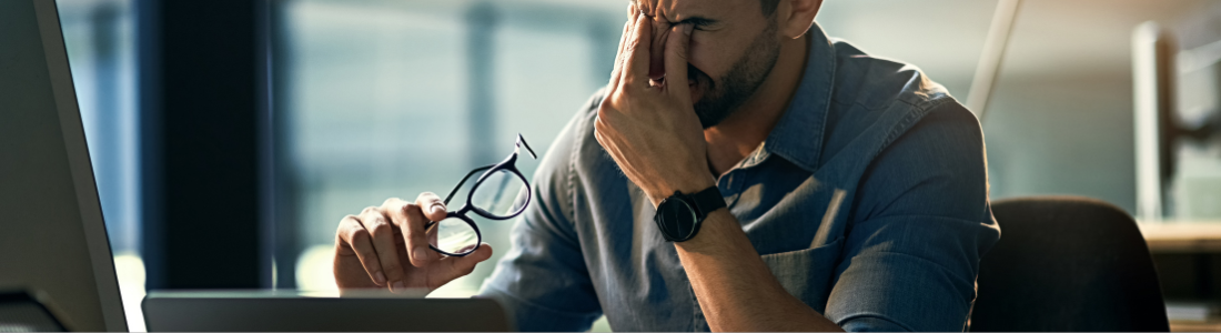 man in front of laptop pinching the bridge of his nose with glasses in the other hand